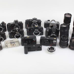 Puede incluir: Una colección de cámaras y lentes Nikon negras. El surtido incluye varios modelos de cámara, lentes de diferentes tamaños y una cámara plateada. Los artículos están dispuestos sobre una superficie blanca.