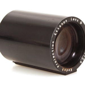 May include: A black Ektanon projection lens with a 4-inch focal length and f/2.8 aperture. The lens has the Kodak brand name and is designed for projection purposes. The lens is cylindrical with a flat base.