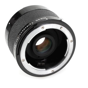 Könnte beinhalten: Ein schwarzes Nikon Telekonverter-Objektiv mit silbernen Akzenten. Das Objektiv hat eine zylindrische Form mit strukturiertem Griff und Objektivöffnung. Das Nikon-Logo ist sichtbar. Für die Fotografie.