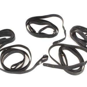 May include: Several black leather straps are arranged on a white background. The straps vary in length and are coiled or looped. Some straps have metal snap closures, suggesting they may be used for securing items.