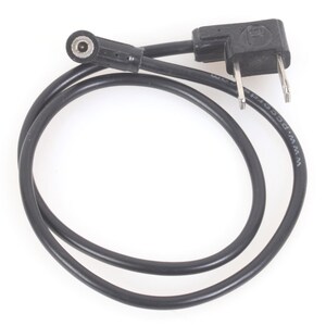 Puede incluir: Cable eléctrico negro con enchufe de dos clavijas y un pequeño conector. El cable está enrollado y descansa sobre una superficie blanca. El enchufe tiene forma rectangular y el conector es circular.
