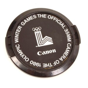 May include: Black Canon 35mm camera lens cap. The cap features white text that reads "1980 Winter Olympic Games The Official 35mm Camera of the Canon". The Olympic rings logo is also visible.