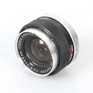 Può includere: Un obiettivo per fotocamera argentato e nero con il testo "TOKYO KOGAKU" e "UV TOPCOR 1:1.8 55 mm" stampato sul lato.