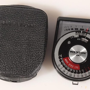 May include: A vintage Weston XM1 light meter with a black leather case. The meter has a circular dial with numbers and settings for film speed and exposure. The case is textured and has a zipper.