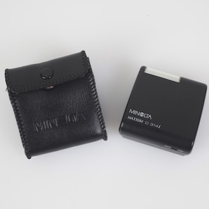 May include: A black Minolta Maxxum D 314iL flash unit and a matching black leather case. The flash unit has a silver top and an on/off switch. The case is embossed with the Minolta logo.
