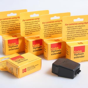 Peut inclure: Plusieurs boîtes Kodak flipflash extender, jaunes et rouges, sont présentées. Elles affichent le texte "Kodak flipflash extender" et des informations sur la réduction des reflets rouges. Un flash noir est également visible.