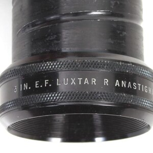 May include: Close-up of a black camera lens with the text "5 IN. E.F. LUXTAR R ANASTIGMAT" in white. The lens has a textured grip and a threaded opening. The lens is a photography accessory.