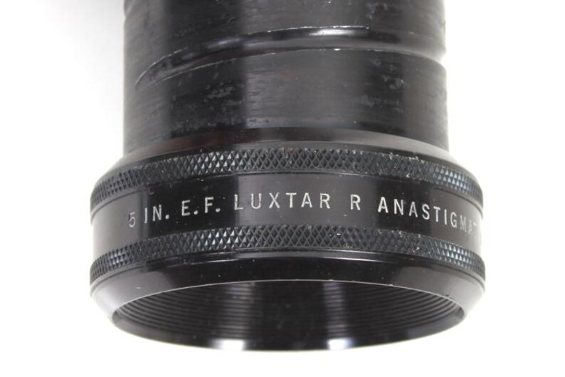 May include: Close-up of a black camera lens with the text "5 IN. E.F. LUXTAR R ANASTIGMAT" in white. The lens has a textured grip and a threaded opening. The lens is a photography accessory.
