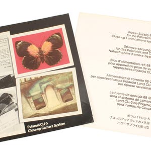 Op de afbeelding: Een zwart-witte Polaroid CU-5 Close-up Camera System doos met een vlinder afbeelding en een wit papier met tekst. De vlinder is bruin en oranje op een roze achtergrond. De tekst is in meerdere talen.