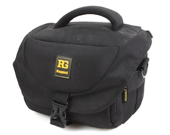 May include: Black camera bag with a yellow and black logo that says "RG Rugged", a black buckle, and the word "JOURNEY" printed on the side.