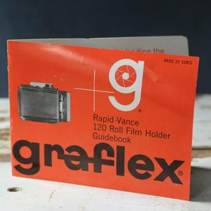 Könnte beinhalten: Ein leuchtend orangefarbenes, gefaltetes Papierhandbuch für einen Graflex Rapid-Vance 120 Rollfilmhalter. Das Cover zeigt eine schwarz-weiße Abbildung des Filmhalters, ein weißes Kamera-Blendenlogo und den Text Graflex in fetten schwarzen Buchstaben.