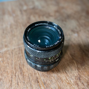 Puede incluir: Un objetivo de cámara negro y plateado con el texto "Soligor 1:2.8 Auto" y "No. 2804803" impreso en el barril del objetivo.