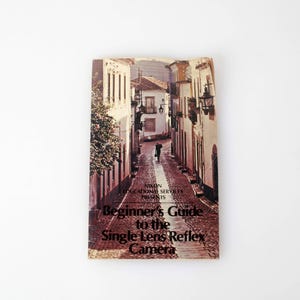 Puede incluir: Un libro antiguo titulado "Beginner's Guide to the Single Lens Reflex Camera". La portada presenta una escena callejera con edificios y una persona caminando por un camino adoquinado. El libro está sobre un fondo blanco.