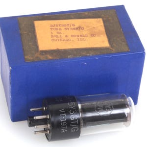 May include: A vintage vacuum tube with a glass enclosure and black base, labelled "-5Y3GT/G VT-197A". It is next to a blue box with a label that reads "3J5T30T/G TUBE SYR3T/G 1 SA. BELL & HOWELL CO. CHICAGO, ILL."
