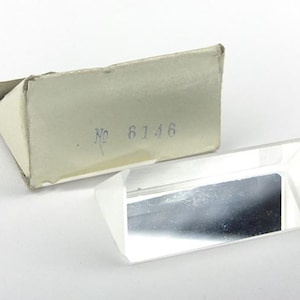 Puede incluir: Un prisma de vidrio transparente con una superficie reflectante. El prisma está empaquetado en una caja de cartón blanca con el número "6146" impreso en ella.