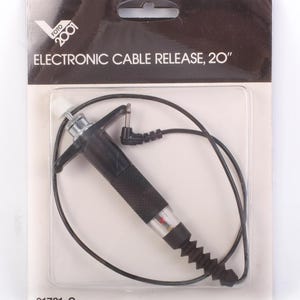 May include: An electronic cable release, 20 inches long, packaged in a clear plastic blister pack. The release is black with a textured grip and a red indicator light. The packaging includes the text "FOTO 2001" and "ELECTRONIC CABLE RELEASE, 20".