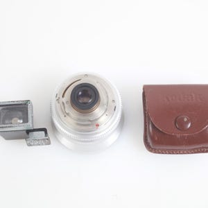Pu&ograve; includere: Un obiettivo per fotocamera vintage, un mirino e una custodia Kodak in pelle marrone. L'obiettivo &egrave; argentato con un anello di apertura nero. Il mirino &egrave; grigio. La custodia Kodak ha una chiusura a scatto.