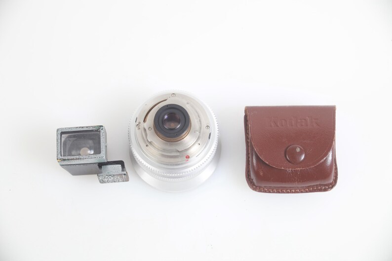 Pu&ograve; includere: Un obiettivo per fotocamera vintage, un mirino e una custodia Kodak in pelle marrone. L'obiettivo &egrave; argentato con un anello di apertura nero. Il mirino &egrave; grigio. La custodia Kodak ha una chiusura a scatto.