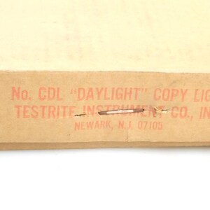 Könnte beinhalten: Nahaufnahme einer Pappschachtel mit rotem Text, der lautet: "No. CDL 'DAYLIGHT' COPY LIGHT TESTRITE INSTRUMENT CO., INC. NEWARK, N.J. 07105". Die Schachtel ist beige und hat Heftklammern und Risse.
