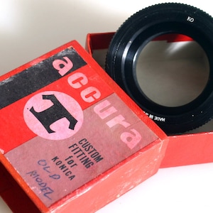 May include: A red box with the words "Accura Custom Fitting for Konica Ol' P Model" printed on it. The box contains a black metal lens adapter ring with the words "Made in Japan" and "KO" printed on it.