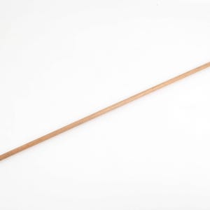 May include: A long, slender wooden dowel rod with a pointed end and a small metal ring at the opposite end. The dowel is a light brown color and appears to be made of natural wood. It is set against a white background.