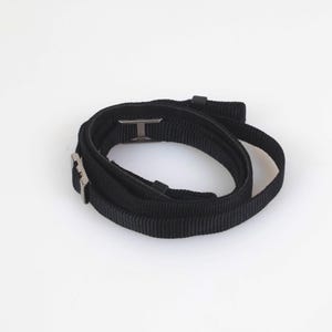 May include: A black nylon webbing strap coiled into a circular shape, featuring a metallic silver-toned rectangular buckle and a sliding adjuster. The strap has a textured, woven surface and is shown against a plain, neutral background.