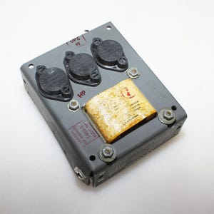 May include: A rectangular gray metal electronic component housing featuring three circular black transistors mounted on the top surface, alongside a rectangular yellowed paper label with printed text and a red logo, secured by four metal bolts.