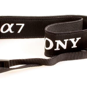 May include: A black camera neck strap featuring the white text SONY and the alpha symbol followed by the number 7. The strap includes a small orange accent detail and black plastic adjustable buckles on the thin nylon webbing ends.