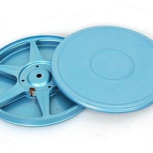 Puede incluir: Un juego de dos carretes de película de metal azul claro. El carrete de la izquierda tiene un cubo central y cinco radios. El carrete de la derecha es una tapa plana.