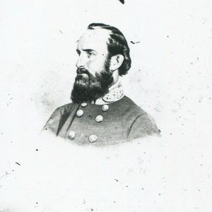 Pode incluir: Um retrato a preto e branco de uma pessoa com barba farta e cabelo escuro, vista de perfil. O indivíduo veste um casaco de estilo militar com uma gola alta com detalhes bordados e duas filas de botões metálicos na parte da frente.