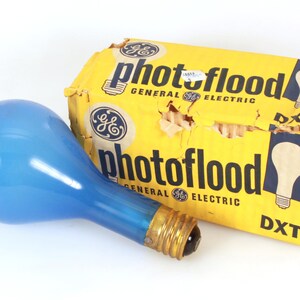 May include: A box of General Electric Photoflood light bulbs. The box is yellow with black text and a blue and white image of a light bulb. A blue light bulb is lying on the box.