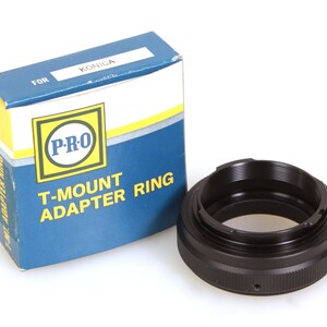 May include: A black T-mount adapter ring for Konica cameras. The ring is packaged in a blue and yellow box with the words "P-RO T-MOUNT ADAPTER RING" printed on it.