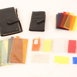 May include: A set of coloured filters for photography, including a variety of colours like red, orange, yellow, green, blue, and purple. The filters are organised in two black leather cases.
