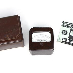 May include: A brown leather case containing a black and white General Electric C/C Light Meter. The meter features a dial with a needle and the words "General Electric" printed on it. The case has a snap closure and a metal button on the front. Instructions for using the meter are included.