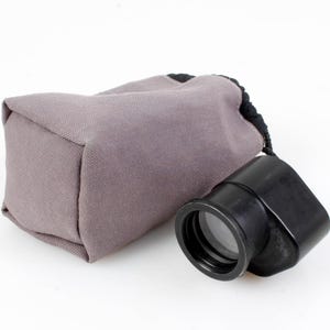 May include: A black loupe with a lens, next to a gray fabric pouch with a black drawstring. The loupe is a magnifying tool, likely for examining small details. The pouch is a protective case.