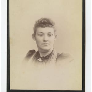 May include: A sepia-toned vintage cabinet card photograph featuring a portrait of a person with curly hair, wearing a dark high-collared garment with lace trim and a thin choker necklace. The bottom border displays the text Smith, S, and Sanford ME.