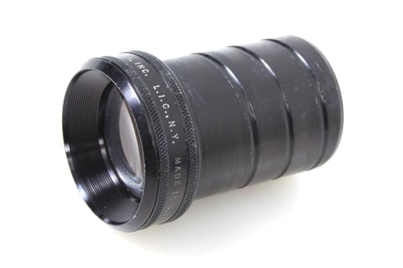 May include: A black camera lens with a ridged body and a lens opening. The lens has the text "L.I.C., N.Y. MADE IN U.S.A." printed on the side. The lens is set against a white background.