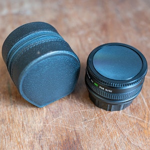 May include: A black camera lens with a black leather case. The lens has a silver band with the text "MC for N/AI" on it.