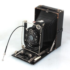 May include: A black vintage folding camera with a bellows system and a lens with a silver rim. The camera has a black leather exterior and a silver metal focusing knob.