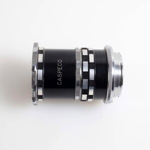 Può includere: Un obiettivo per fotocamera nero e argento con la scritta "CASPECO" stampata sul lato. L'obiettivo ha una forma cilindrica con anelli e dettagli argentati. L'obiettivo è posto su uno sfondo bianco.