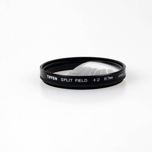 May include: A black circular Tiffen Split Field +2 67mm camera lens filter. The filter features a clear glass element that covers half of the circular frame, with white text printed on the side indicating the brand, type, power, and size.