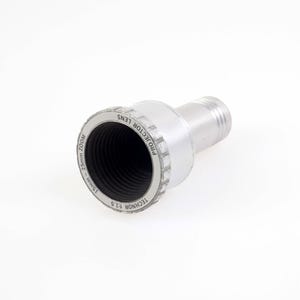 Op de afbeelding: Zilveren projectorlens met een zwart interieur. De lens heeft de tekst "PROJECTOR LENS" en "TECHNOR F1.5" op de buitenste ring. De lens is ontworpen voor een 25 mm zoom en een 15 mm zoom.
