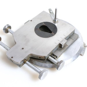 May include: A silver metal device with a circular base and a rotating platform. The platform has a circular opening with a black centre and a small, triangular cutout. The device has two knobs on the side and a scale marked in degrees.