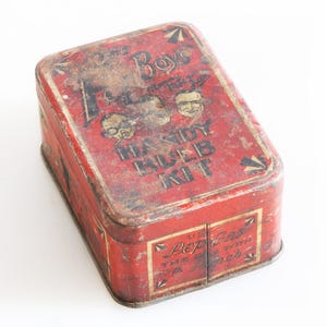 May include: A rectangular, vintage red metal tin, featuring the text "The Boys Handy Rub Kit" and "Pep Gas with a Punch." The tin has a hinged lid and shows signs of wear and age, with a distressed appearance.
