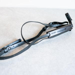 Puede incluir: Correa de cámara de cuero negro con una hebilla de metal y un conector de correa de cámara de plástico negro con la palabra "camera" impresa en él.