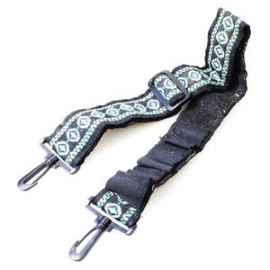 Op de afbeelding: Zwarte schouderriem met decoratief patroon en metalen sluitingen. Het patroon is wit, turquoise en geel. Het heeft een zwarte kunststof gesp en metalen sluitingen om aan een tas te bevestigen. De riem is ontworpen voor een tas of handtas.