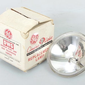 May include: A white box with red text that reads "This package contains one DWG Narrow Beam Lamp for Telephoto Lenses. Manufactured by General Electric Cinema Light."  The box is next to a clear glass replacement lamp for a Newport Battery Portable Cinema Light.