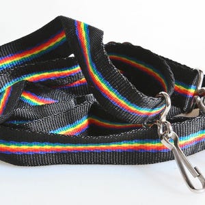 May include: Black nylon strap with a rainbow stripe design. The strap has two silver-colored metal swivel clips. The rainbow stripe includes red, orange, yellow, green, blue, and purple colors. The strap is likely for a bag or pet leash.