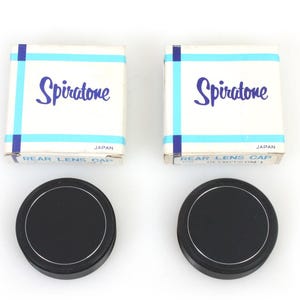 Puede incluir: Dos tapas traseras de lente negras con borde plateado, junto a sus cajas originales. Las cajas son blancas con detalles azules y la palabra "Spiratone" en cursiva. Las cajas también dicen "Rear Lens Cap" y "Japan".