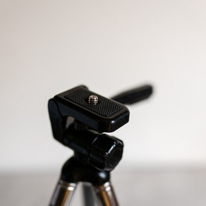 Puede incluir: Un tr&iacute;pode negro con una cabeza texturizada y ajustable. La cabeza tiene un tornillo para sujetar una c&aacute;mara u otro dispositivo.